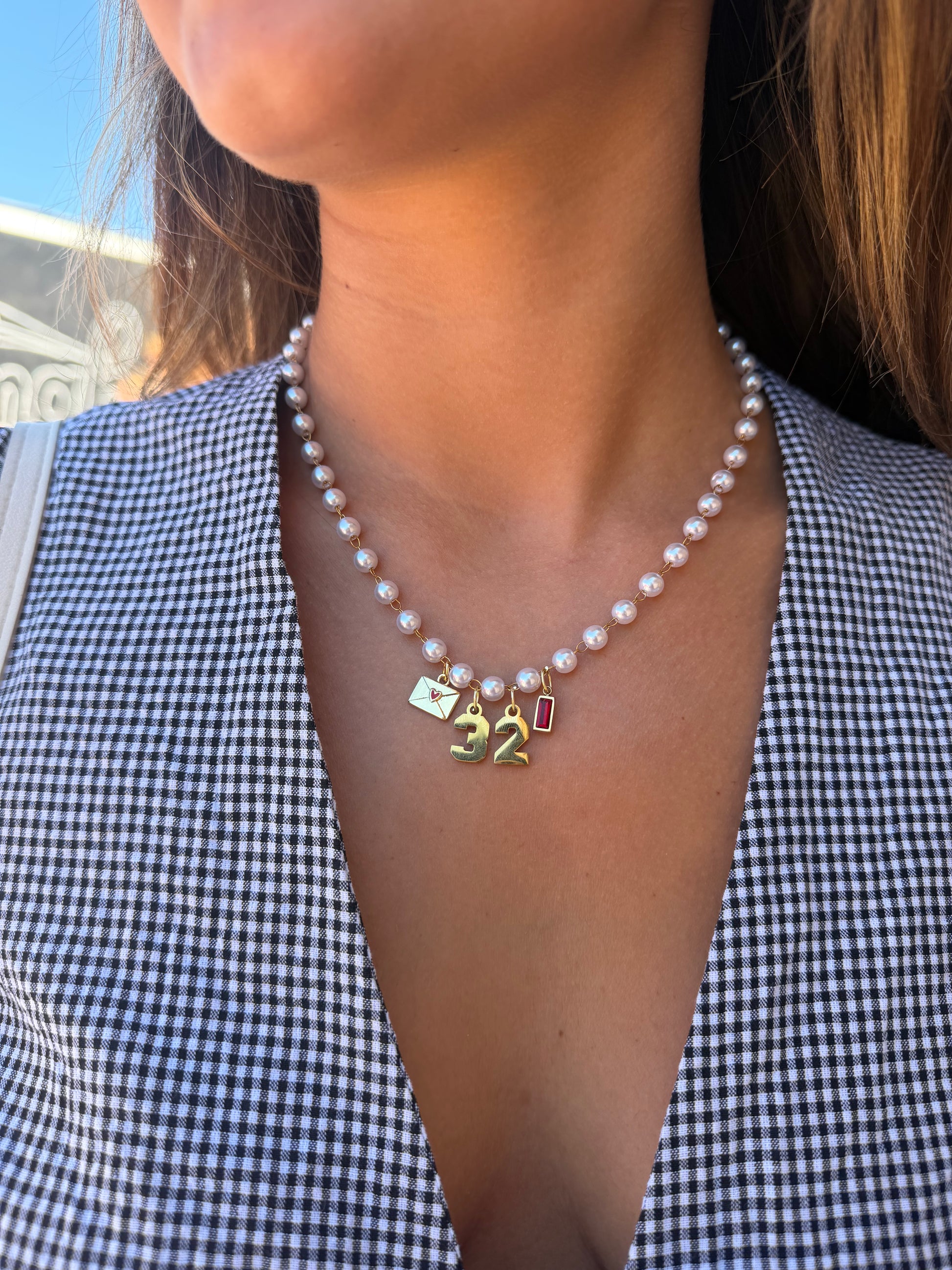 A close-up of a woman wearing a pearl charm necklace with gold-toned charms, including a '32' number charm and a small rectangular charm, paired with a black and white gingham top.
