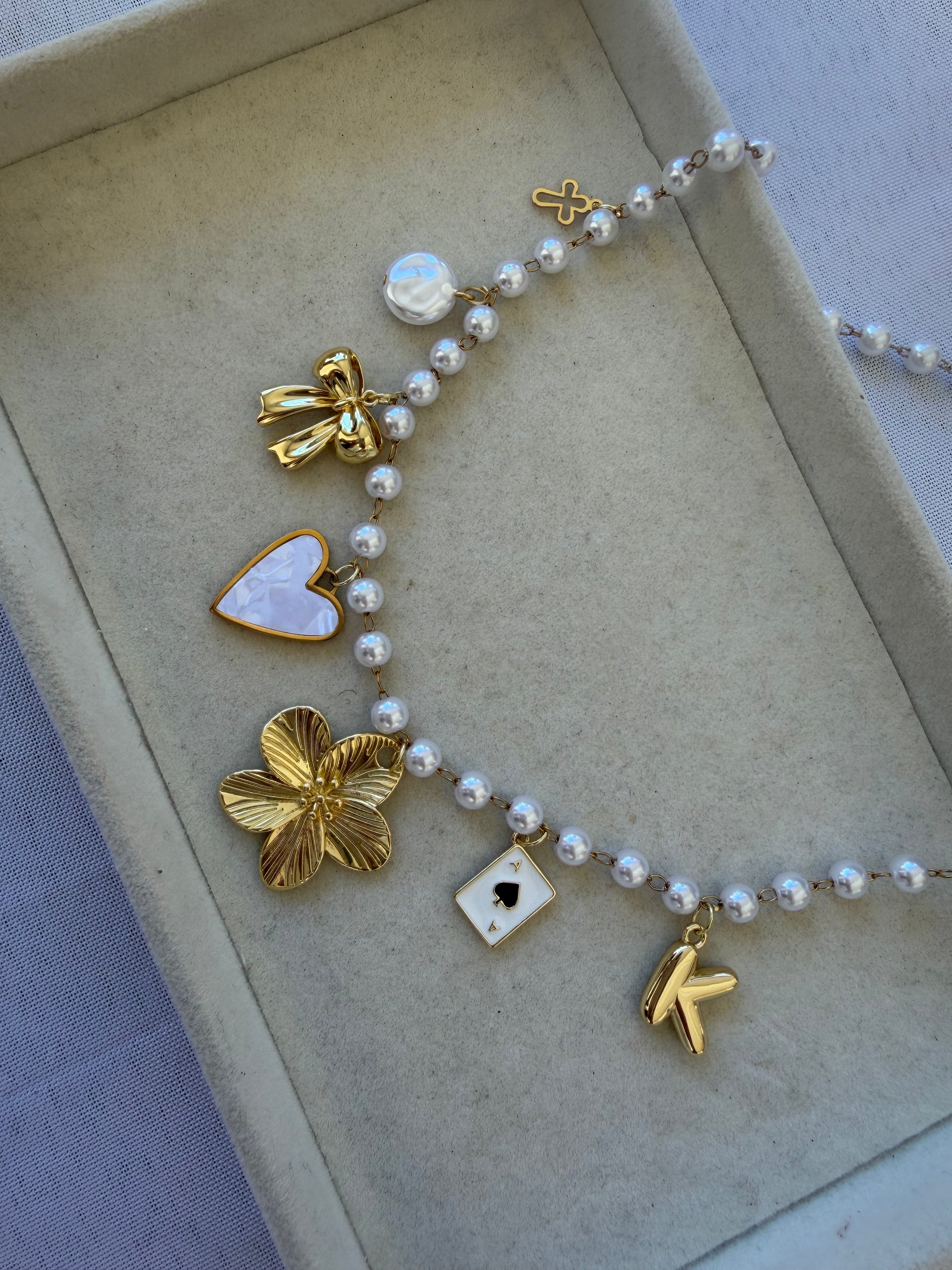 A pearl and gold charm necklace displayed inside a beige jewelry box. The necklace features white pearls threaded with gold-toned charms, including a bow, a heart-shaped moonstone, a flower, a playing card, and a bird.
