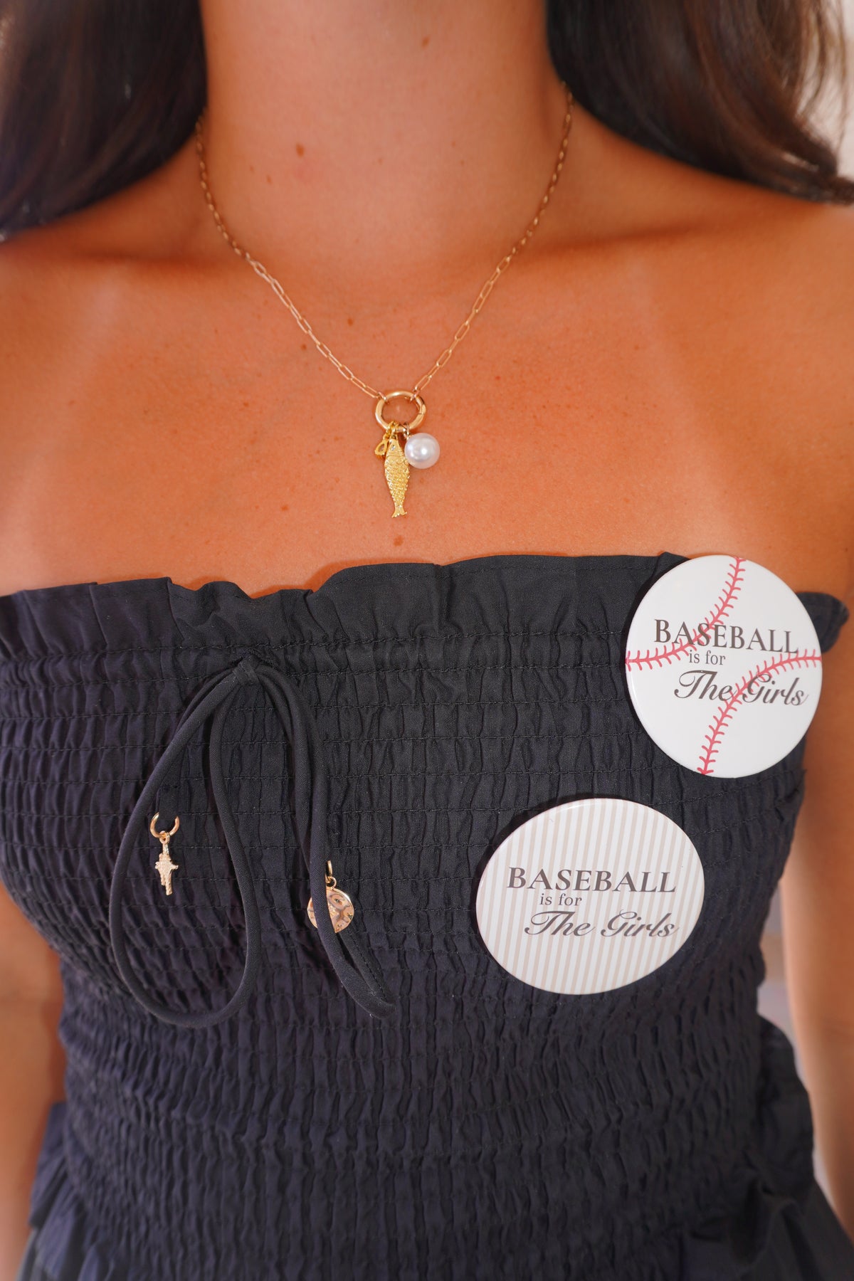 Close-up of a woman wearing a gold necklace with a baseball-themed charm, a black smocked top, and two round white pins that read 'BASEBALL FOR THE GIRLS'.