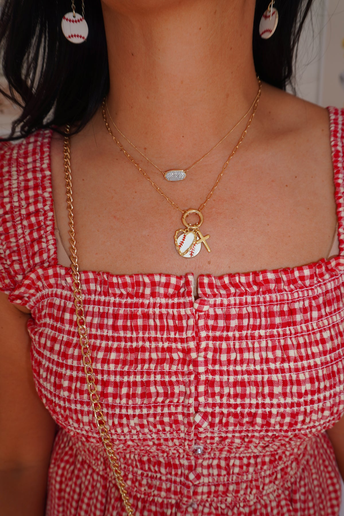 A close-up lifestyle shot of a person wearing a gold double-layered necklace with a baseball pendant and a small tennis ball charm, paired with a red and white gingham top.