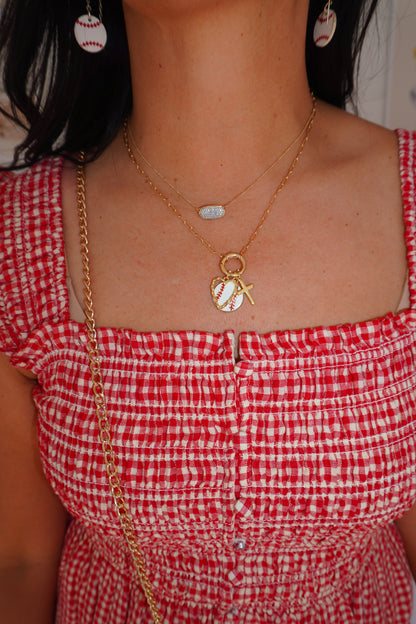 A close-up lifestyle shot of a person wearing a gold double-layered necklace with a baseball pendant and a small tennis ball charm, paired with a red and white gingham top.